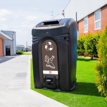Un gran armario para contenedor de residuos alimentarios, dise&ntilde;ado para alojar un contenedor con ruedas en un espacio p&uacute;blico, rodeado de c&eacute;sped verde y situado cerca de un sendero peatonal.
