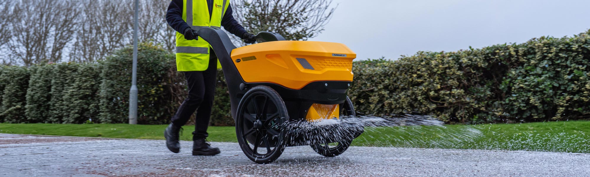 Un &eacute;pandeur manuel de sel et de gravillons utilis&eacute; sur un parking en hiver par une personne portant une veste haute visibilit&eacute;.