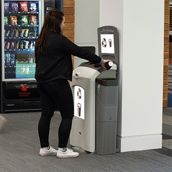 Une station de recyclage Nexus 100 pour gobelets et liquides, plac&eacute;e contre un pilier blanc dans un caf&eacute; ou un bistro public, tandis qu'un passant verse du liquide dans le bac de collecte.