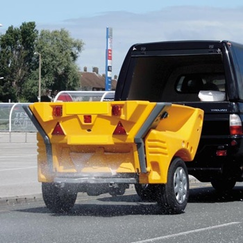 Un &eacute;pandeur remorquable de sel ou de gravillons Glasdon Turbocast 1000, tract&eacute; par une camionnette et r&eacute;pandant du sel sur un parking en hiver.