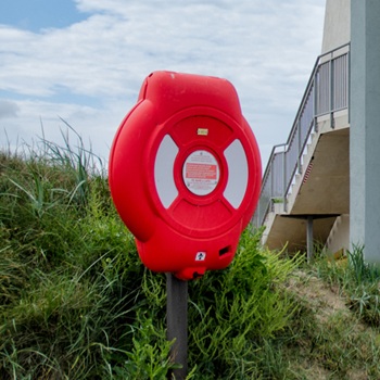 A life ring housing in front of some green foliage, next to a building on a beach