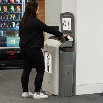 A Nexus 100 cup and liquid recycling station placed against a white pillar in a public caf&eacute; or bistro with a passerby disposing a cup into it