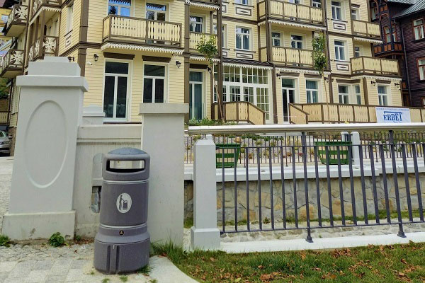 Spersonalizowany kosz na śmieci Mini Plaza ustawiony przy betonowej ścianie z zabudowaniami z tyłu