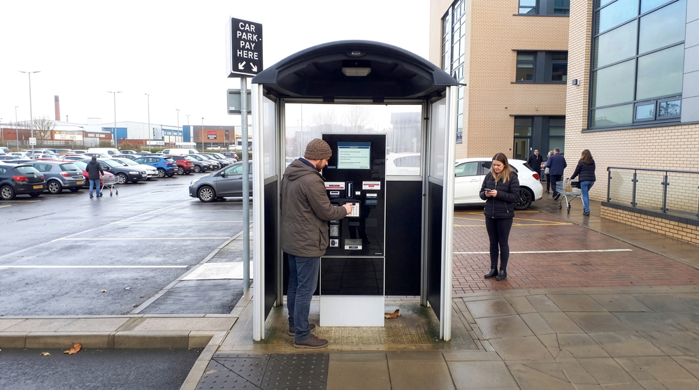 Image of a Modus ticket machine shelter on a car park with people walking by and paying for their parking ticket