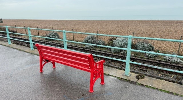 red british heart foundation bench on a promenade overlooking a beach
