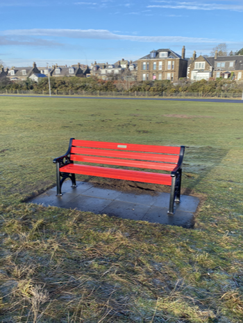 front view of a red british heart foundation bench in a grassland with trees and houses in the background