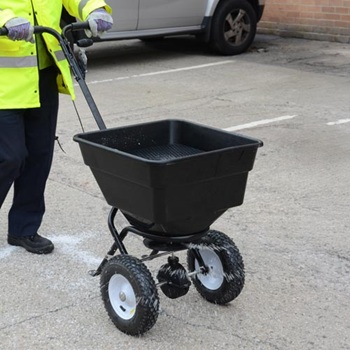 A Glasdon Icemaster manual salt and grit spreader being driven over a car park in winter by a person in a hi-viz jacket.