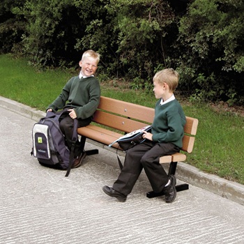 A brown Glasdon bench on a pavement with a green backdrop with two school children sitting on it