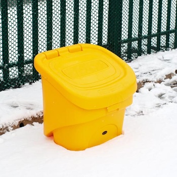 A Glasdon Nestor 90 grit and salt bin in front of a green metal and mesh fence in winter surrounded by snow.