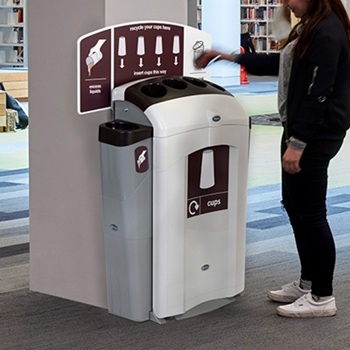 A Nexus 100 cup and liquid recycling station placed against a white pillar in a public caf&eacute; or bistro with a passerby disposing a cup into it