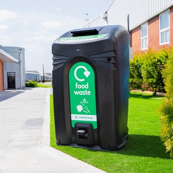 A large food waste bin housing for a wheelie bin in a public space, surrounded by green grass and near a footpath