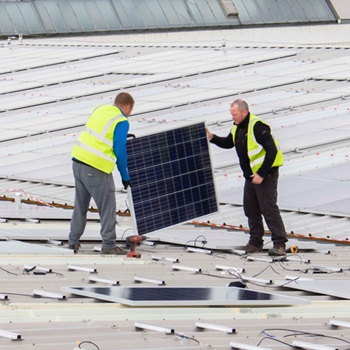 Two people in hi-vis vests on a rooftop ready to place down a large solar panel cell to install it