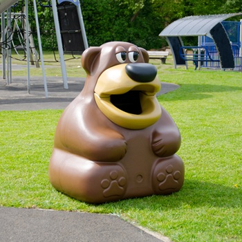 A bear shaped tidybear Glasdon bin on a green field next to a walkway and near a playground on a sunny day
