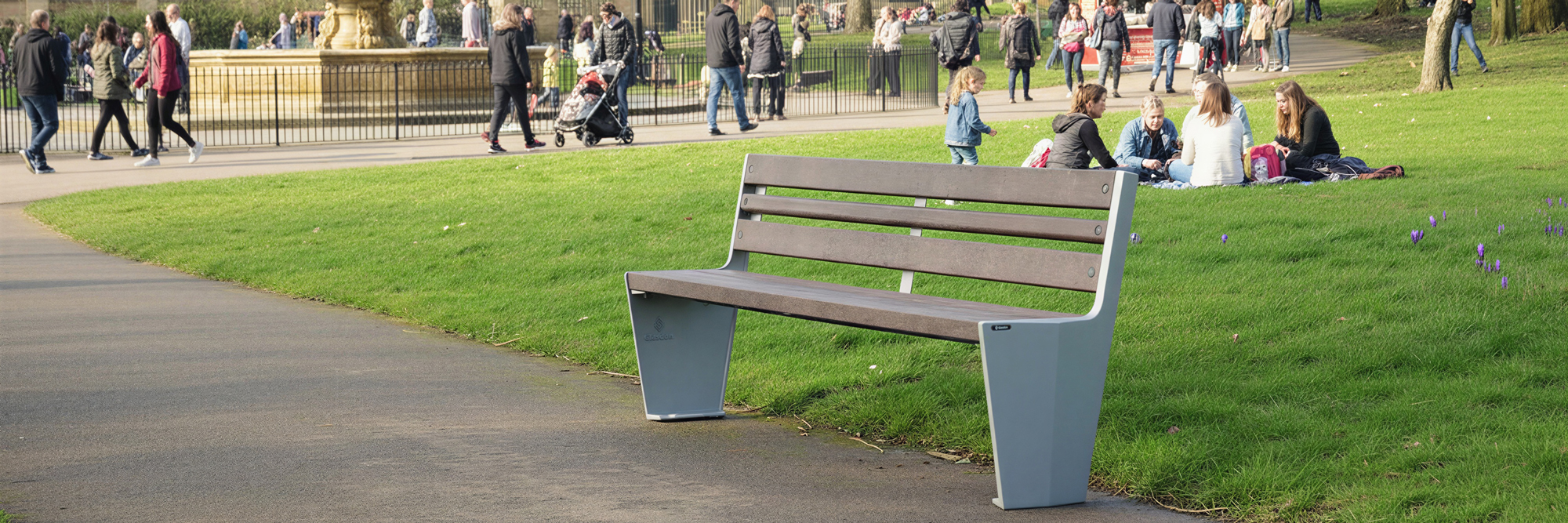 A nice bench in a park with green fields and people sitting down to have a picnic