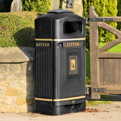 A black Streamline Jubilee Bin standing at the corner of a low stone wall, topped by a green hedge and beside a wooden garden gate