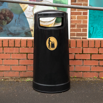 A black Topsy 65 bin stood at the edge of a road with a railing and red brick wall behind it