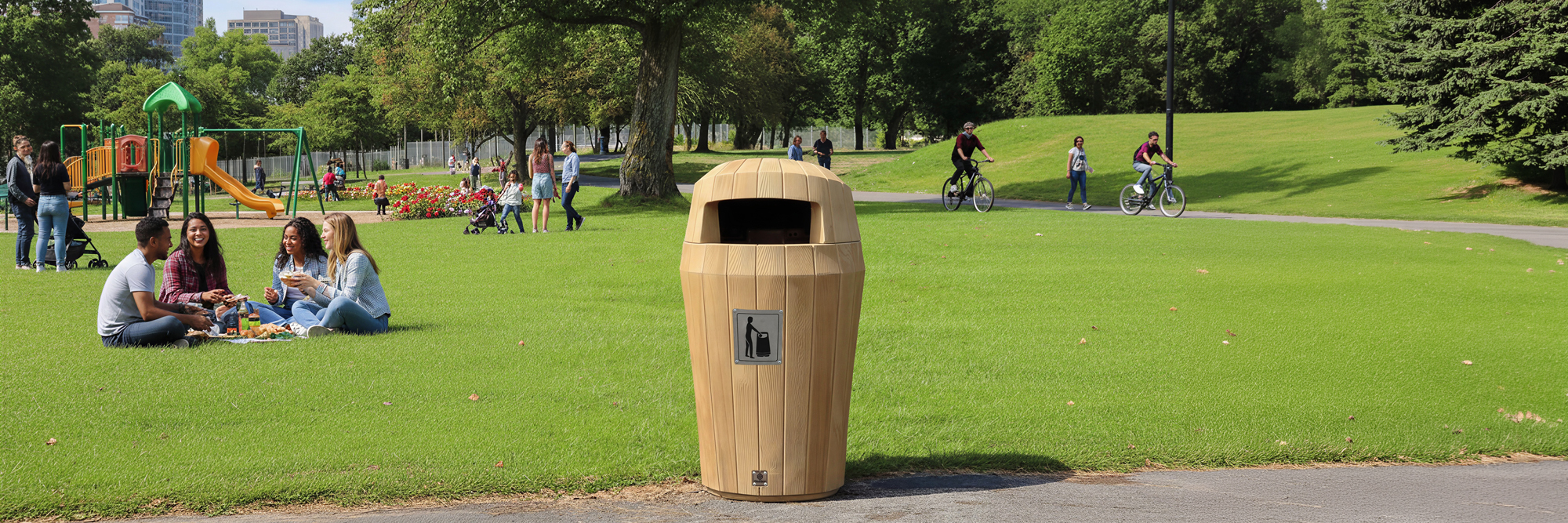 A nice wooden looking garbage can in a park with green fields and people sitting down to have a picnic
